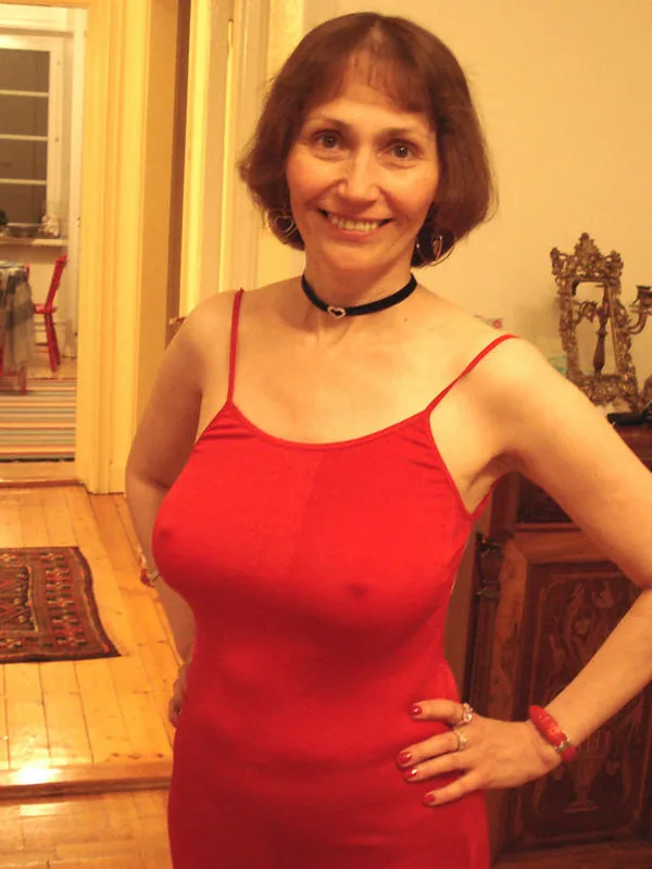 Femme souriante en robe rouge posant dans un intérieur avec parquet et meubles anciens.