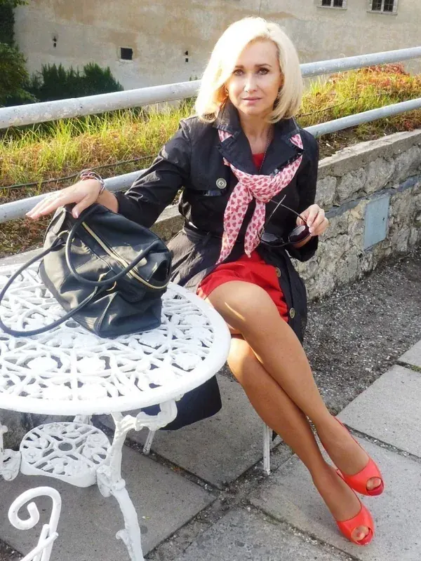 Femme blonde assise à une table blanche en extérieur, portant une jupe rouge, une veste noire et des sandales rouges.
