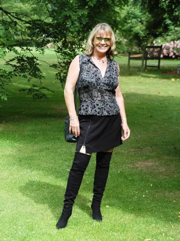 Femme blonde en tenue sombre posant en plein air sur une pelouse, sous un arbre, avec des lunettes de soleil.