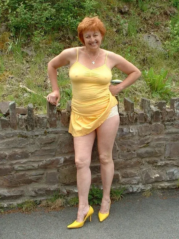 Femme aux cheveux roux en robe jaune et talons, posant devant un mur de pierre en extérieur.