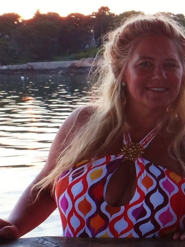 Femme blonde en maillot de bain coloré souriant devant l'eau au coucher du soleil.