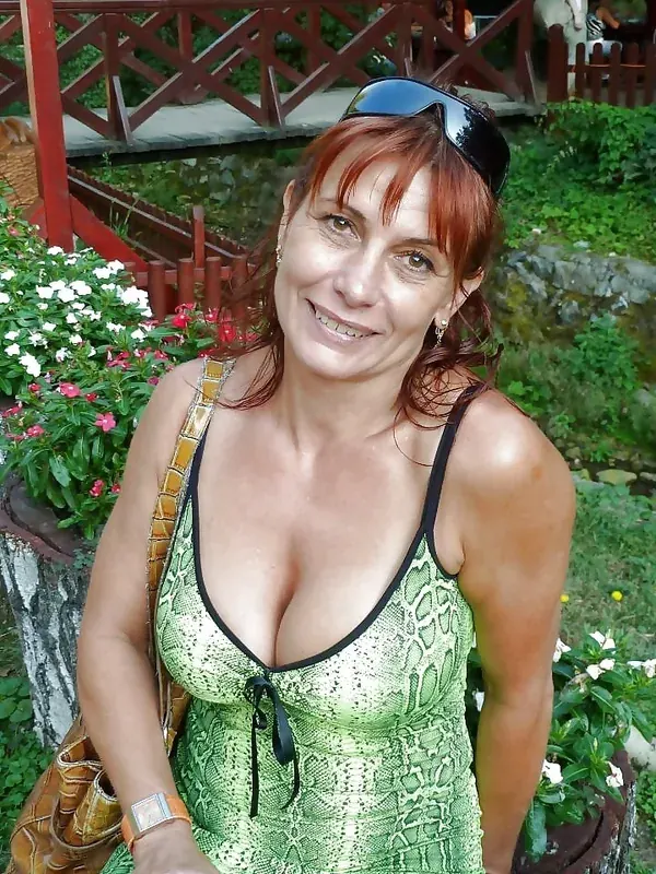 Femme aux cheveux roux en robe verte à motif serpent, souriante, en extérieur près de fleurs et d'une structure en bois.