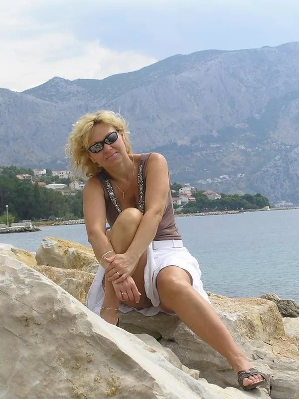 Femme blonde en lunettes de soleil assise sur des rochers près de la mer, avec des montagnes en arrière-plan.