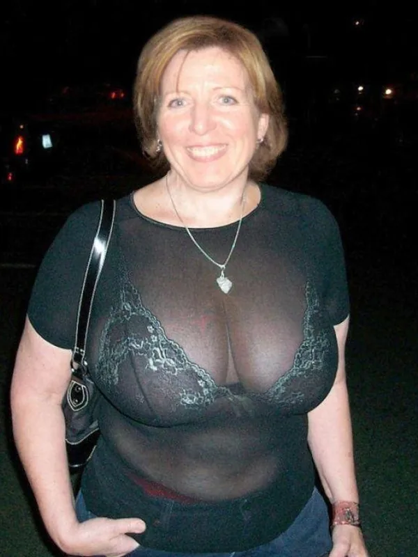 Portrait en buste d'une femme souriante portant un haut noir transparent et un soutien-gorge en dentelle, prise de nuit.