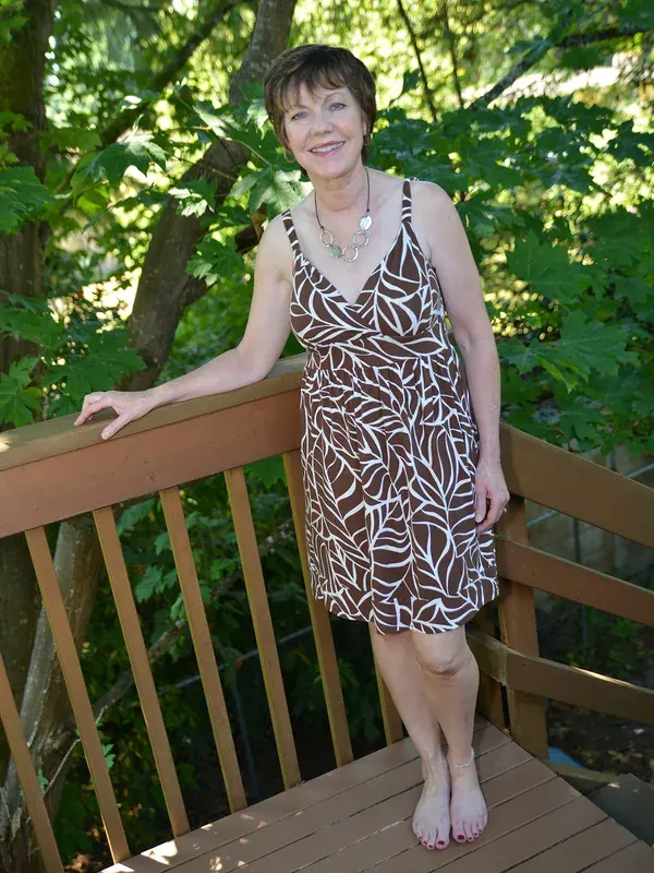 Femme souriante en robe à motifs feuillagés, debout sur une terrasse en bois entourée de verdure.