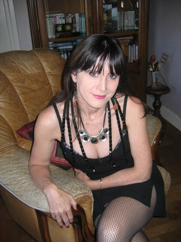 Femme aux cheveux noirs assise sur un fauteuil en cuir, portant une robe noire et des collants résille, dans un salon intérieur.