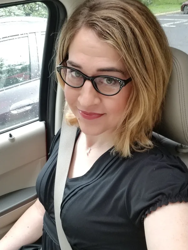 Portrait d'une femme aux cheveux blonds et lunettes, assise dans une voiture, regardant l'objectif, avec une ceinture de sécurité.