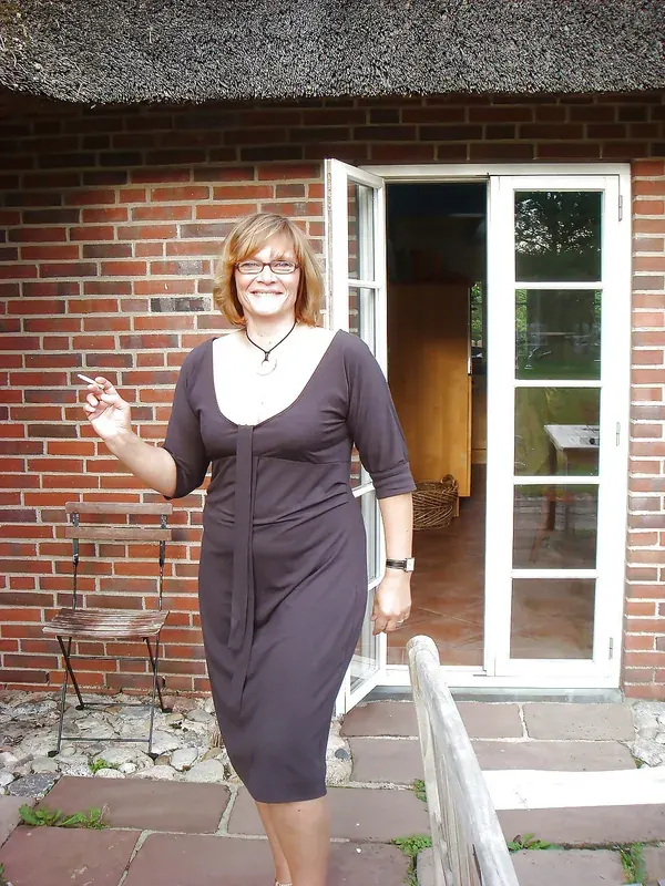 Femme souriante en robe marron tenant une cigarette devant une maison en briques avec toit de chaume.