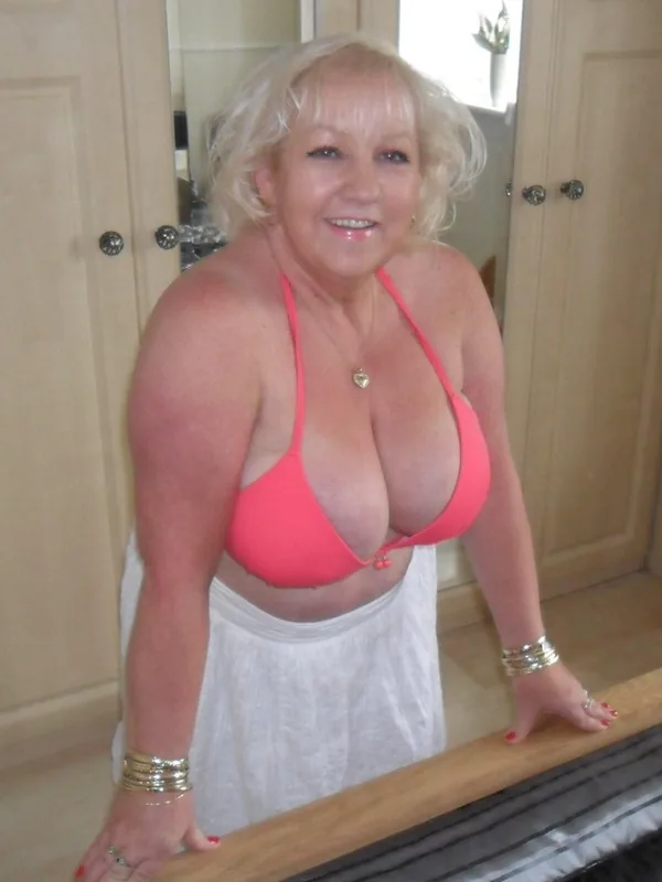 Femme blonde en haut de bikini rose et jupe blanche, appuyée sur un lit dans une chambre.