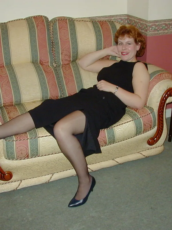 Femme aux cheveux roux assise sur un canapé rayé, portant une robe noire et des chaussures à talons, dans un salon.