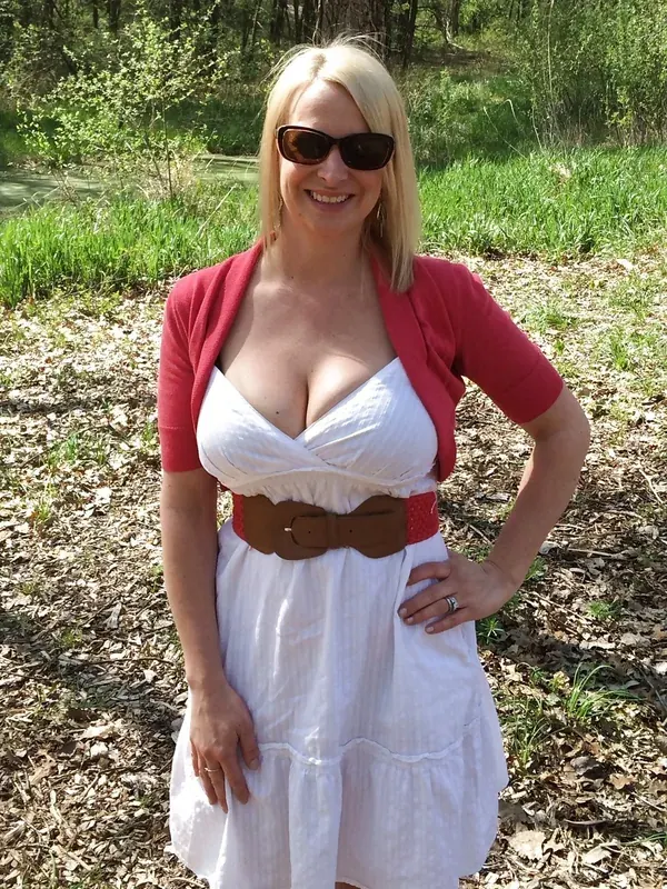 Femme blonde en robe blanche et cardigan rouge, souriante, debout en extérieur sous un arbre.